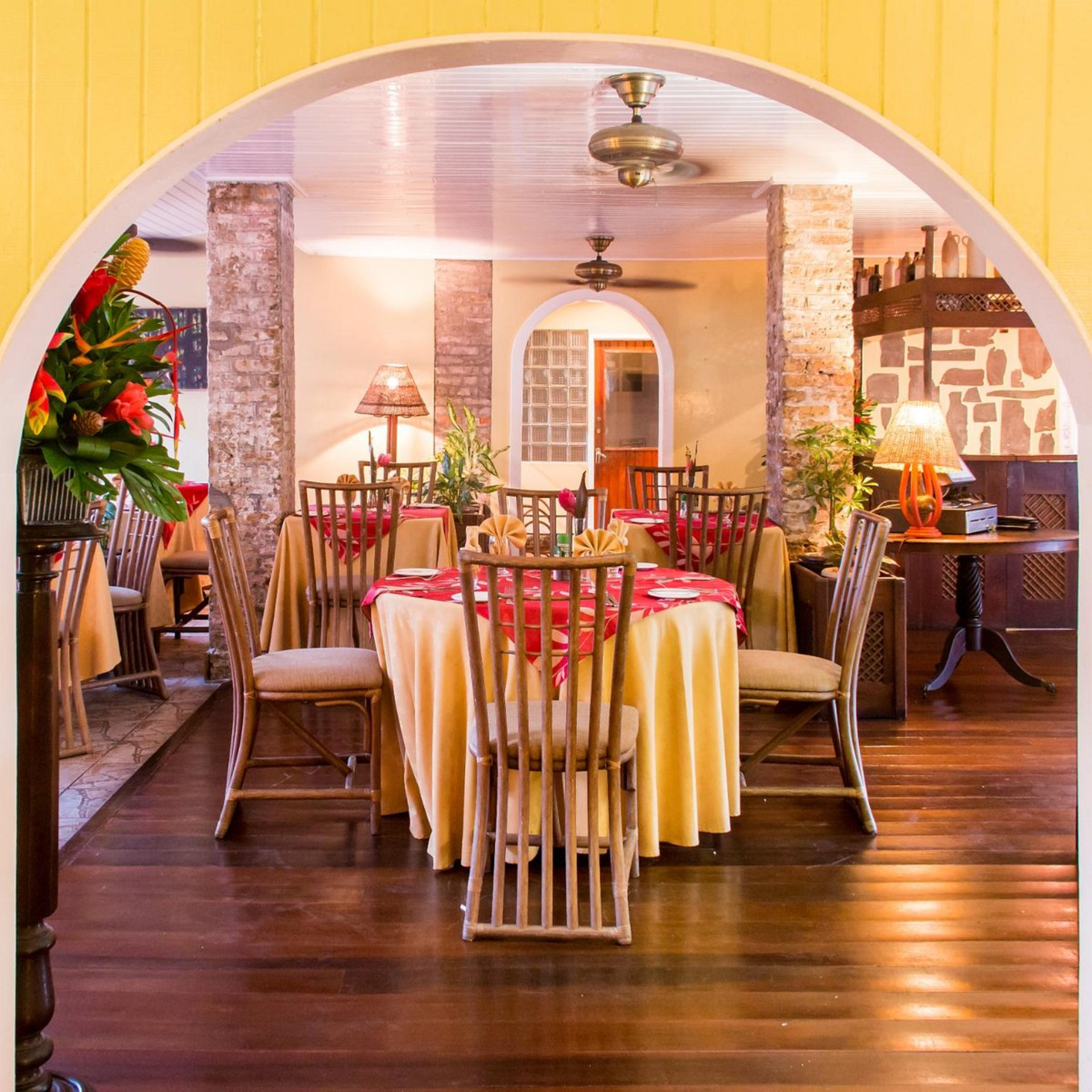 Warmly lit restaurant dining room with yellow tablecloths, red runners, wooden chairs, brick columns, and cozy rustic décor