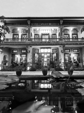 Black and white photo of ornate two‑story building with decorative carvings, tiled roof, courtyard pond, and illuminated windows at night