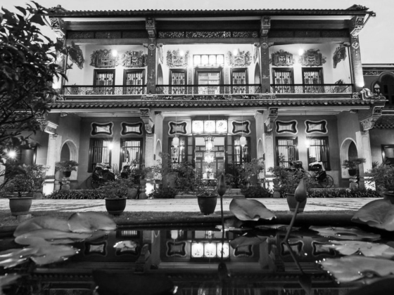 Black and white photo of ornate two‑story building with decorative carvings, tiled roof, courtyard pond, and illuminated windows at night