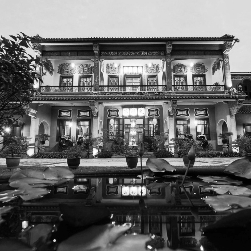 Black and white photo of ornate two‑story building with decorative carvings, tiled roof, courtyard pond, and illuminated windows at night