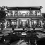 Black and white photo of ornate two‑story building with decorative carvings, tiled roof, courtyard pond, and illuminated windows at night