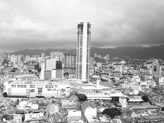 Black and white cityscape with modern skyscraper rising above dense urban buildings and distant mountains under cloudy sky