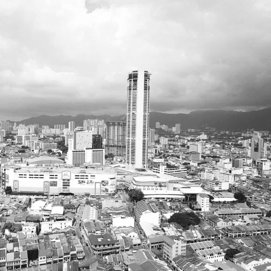 Black and white cityscape with modern skyscraper rising above dense urban buildings and distant mountains under cloudy sky