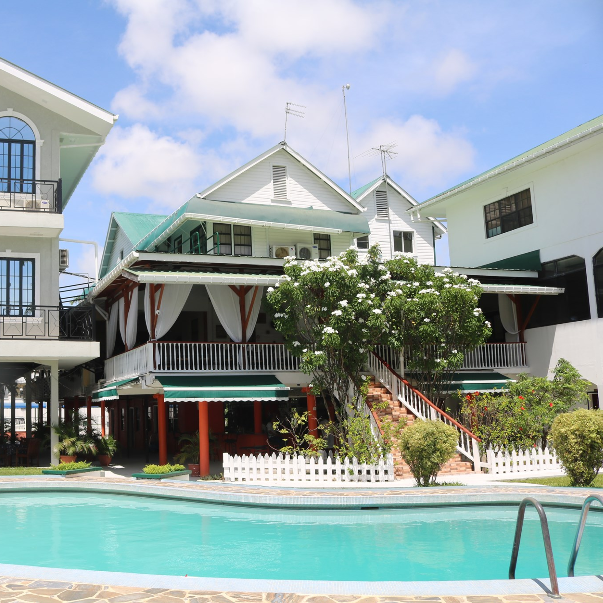 Tropical resort with white buildings, green roofs, veranda with red columns, swimming pool, and lush gardens under a partly cloudy sky