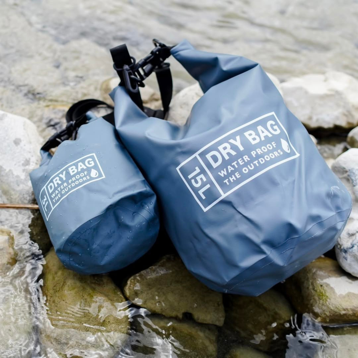 Two blue waterproof dry bags labeled The Outdoors placed on rocks by shallow river stream