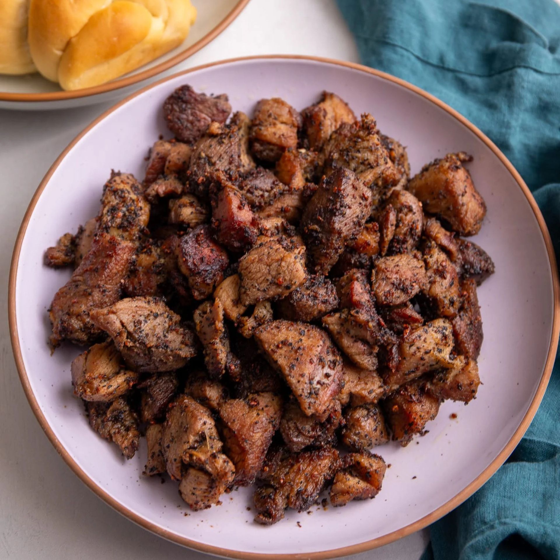 Grilled pepper-crusted meat on plate with bread rolls in background and teal cloth beside